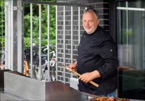 Professionelle Personal Branding Fotografie im Münsterland: Menschenfotograf Thomas Schoo fängt das smarte Lächeln von Gastronom Marco Althoff (Althoff`s Landhaus Langenhorst) mit Grillbesteck haltend in einer entspannten Shooting-Atmosphäre ein.