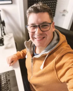 Dieses Foto zeigt Sascha Übler in seinem Office! Er selbst hat dieses Selfie geschossen. Er trägt einen orangebraunen Hoddie, mit grauem Innenfutter – im Ansatz ist seine Tastatur in der Unschärfe zu sehen. Untypisch trägt er seine Arbeitsbrille für den Einsatz von Sicherheit im Business!