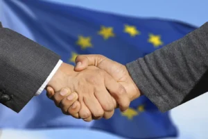 Vertrauen zeigt sich in einer Businesspartnerschaft oft sehr schnell – Sascha Übler Versicherungen und ich gaben uns die Hände auf faire Zusammenarbeit, im Hintergrund ist die Fahne der Europäischen Union in der Unschärfe zu sehen!